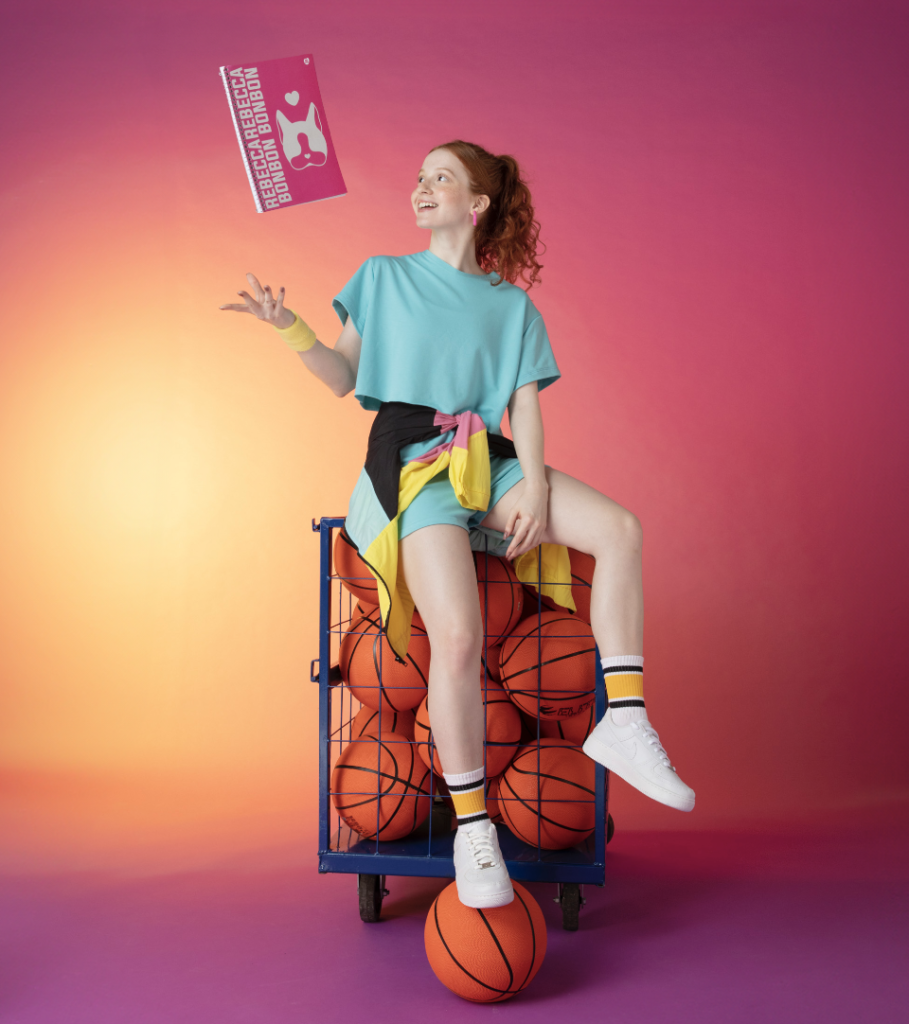 Modelo em uma cesta de basquete, com o caderno da Rebecca Bonbon com a Animativa.